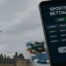 Hand holding smartphone displaying sports betting app with Parliament Hill Peace Tower in background, representing Canada's Bill S-211 advertising regulations